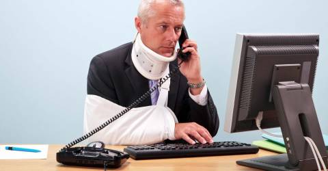 Gewonde zakenman met arm in mitella en nekbrace zit achter zijn computer aan zijn bureau aan de telefoon